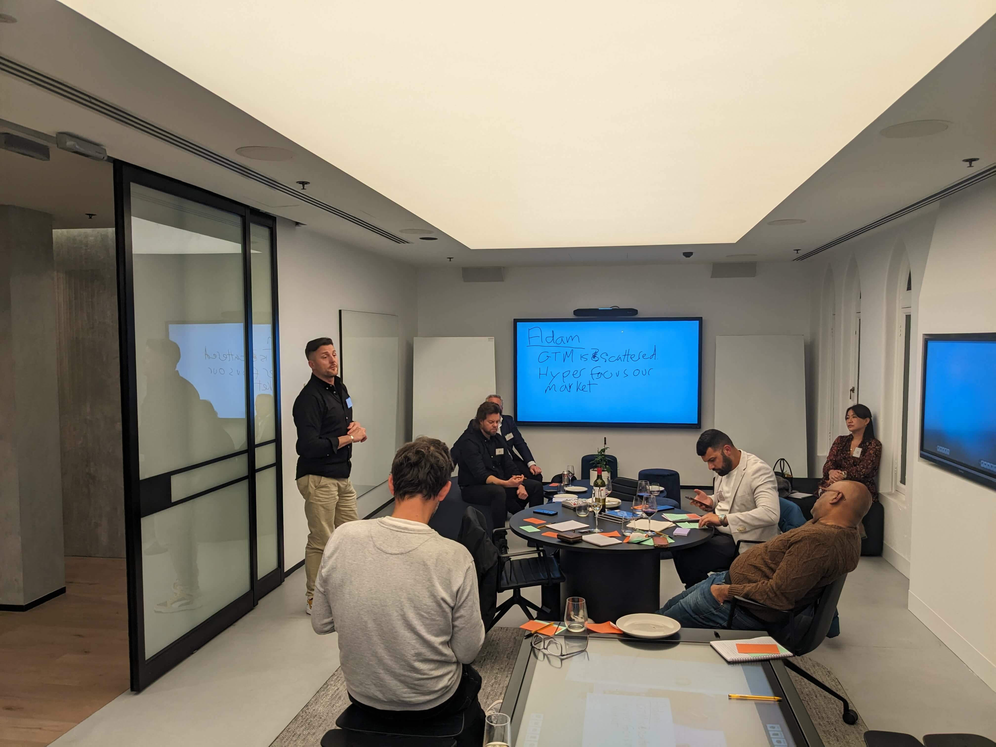 A group of people gather around a table in front of a whiteboard, they have glasses of wine and are exchanging ideas.